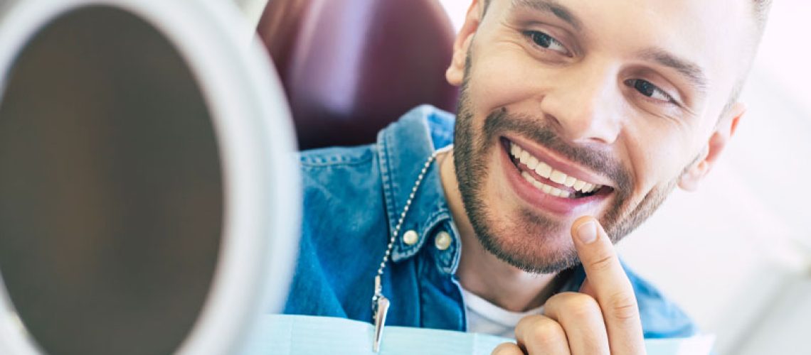 a dental patient smiling into a hand held mirror while pointing to his teeth because he just improved his smile with dental veneers.
